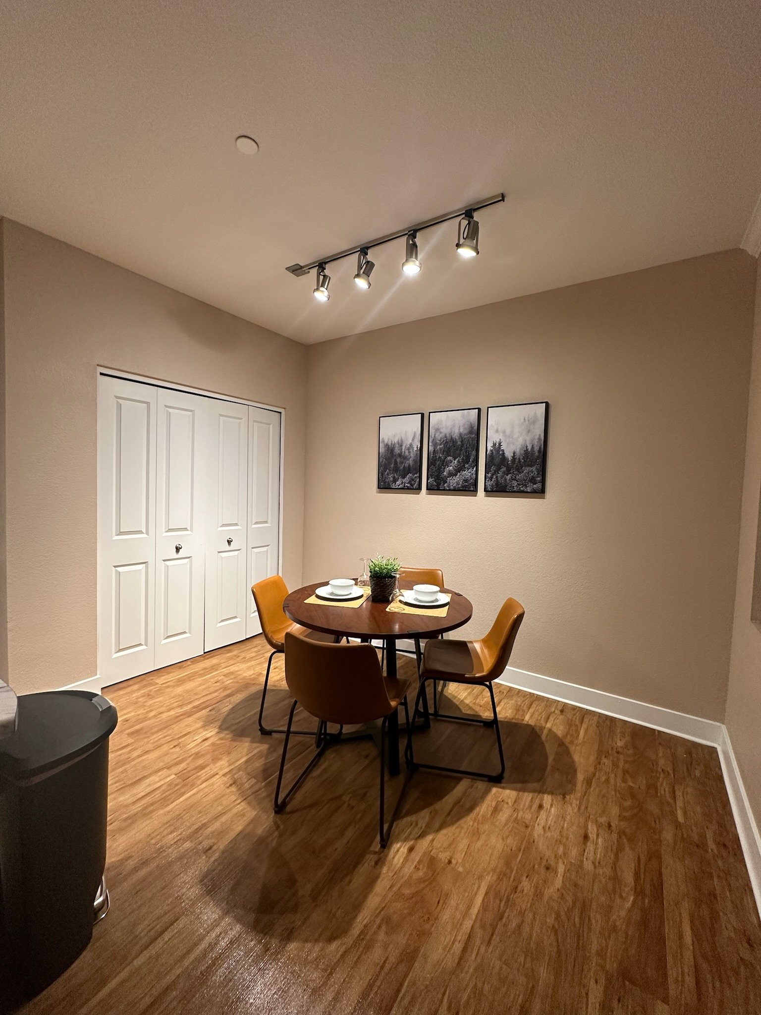 Dining area with a round wooden table set with white plates and cups, four brown chairs, a small plant in the center, three framed black and white landscape photographs on the beige wall, hardwood floors, and a ceiling track light.
