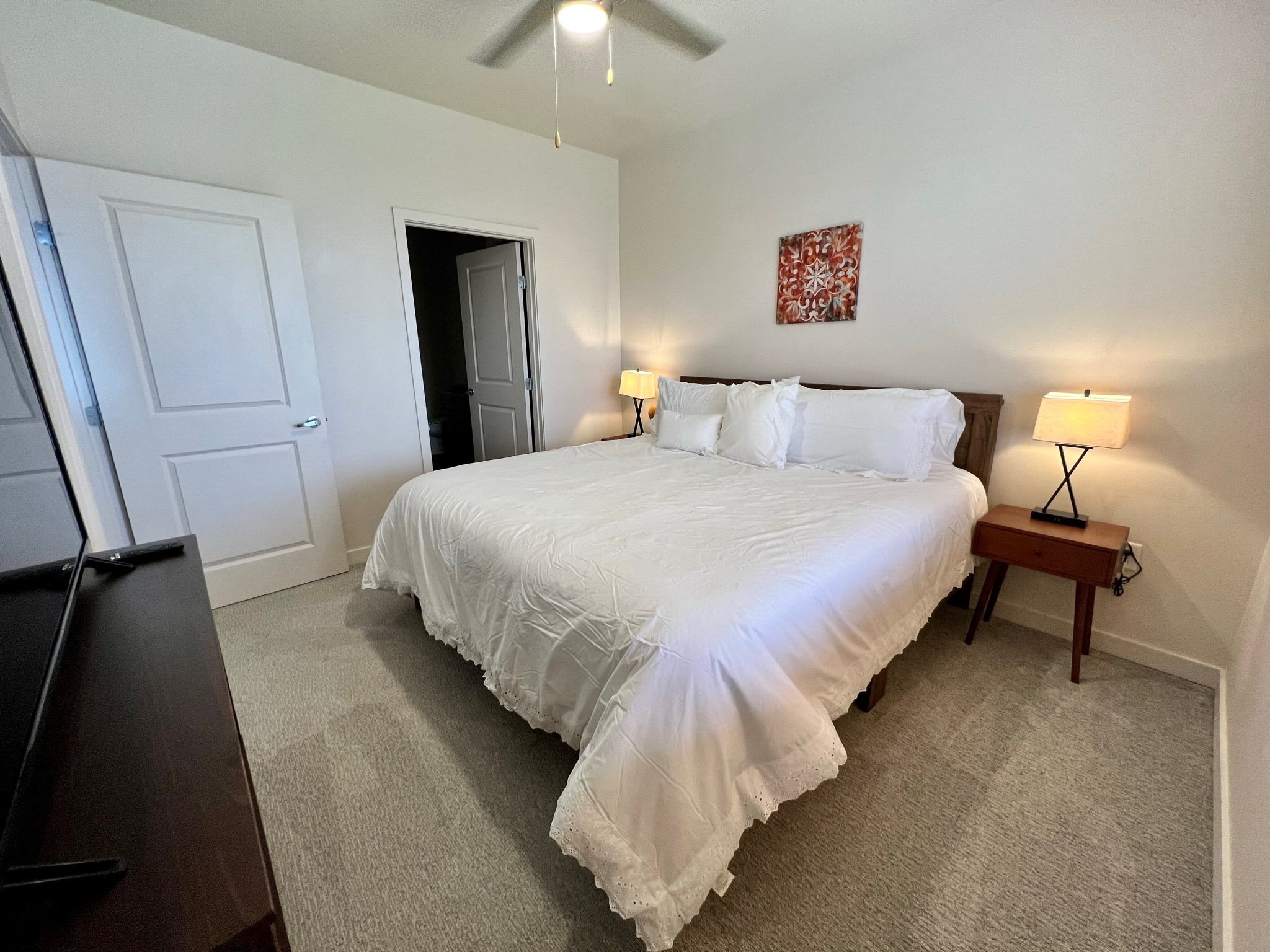 A neatly made bed with white bedding and multiple pillows in a bedroom with beige carpet, side tables with lamps, and a wall art piece above the headboard.