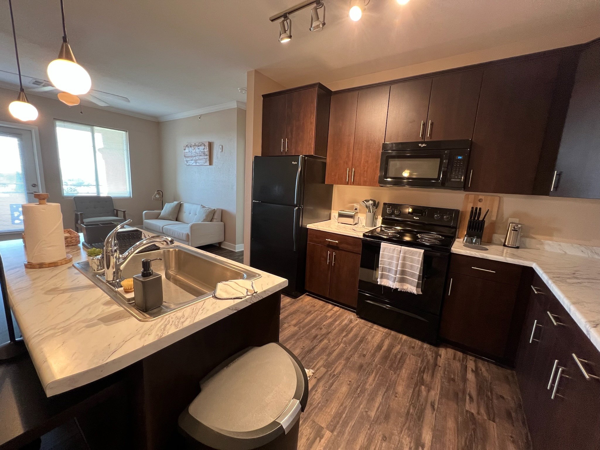 Open-concept kitchen with dark wood cabinets, black appliances, marble countertops, and a living area with a beige sofa and a window with blinds.
