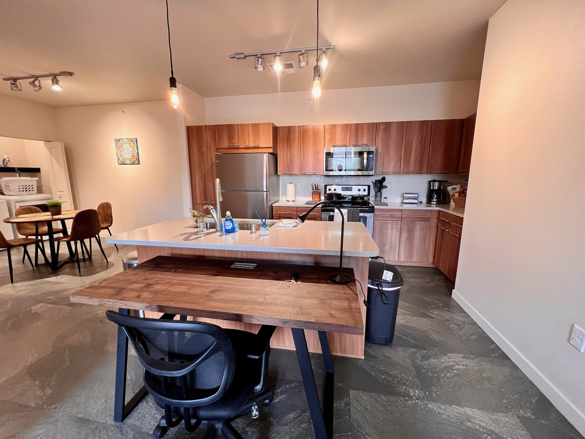 Modern kitchen with wooden cabinets, stainless steel appliances, a white kitchen island with a sink, and a dining area with a round table and six chairs in the background.