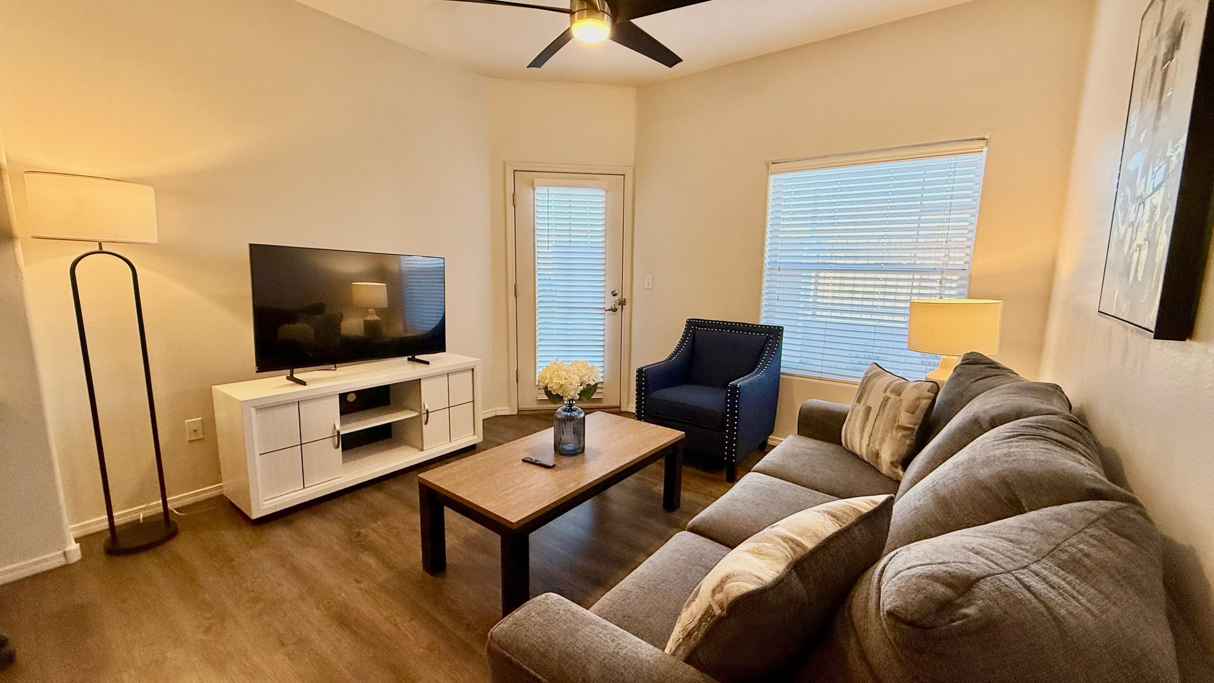 Living room with beige walls, a gray sofa, a black armchair, a wooden coffee table with a vase of white flowers, a white TV stand with a flat-screen TV, a window with blinds, a door with blinds, and a ceiling fan.