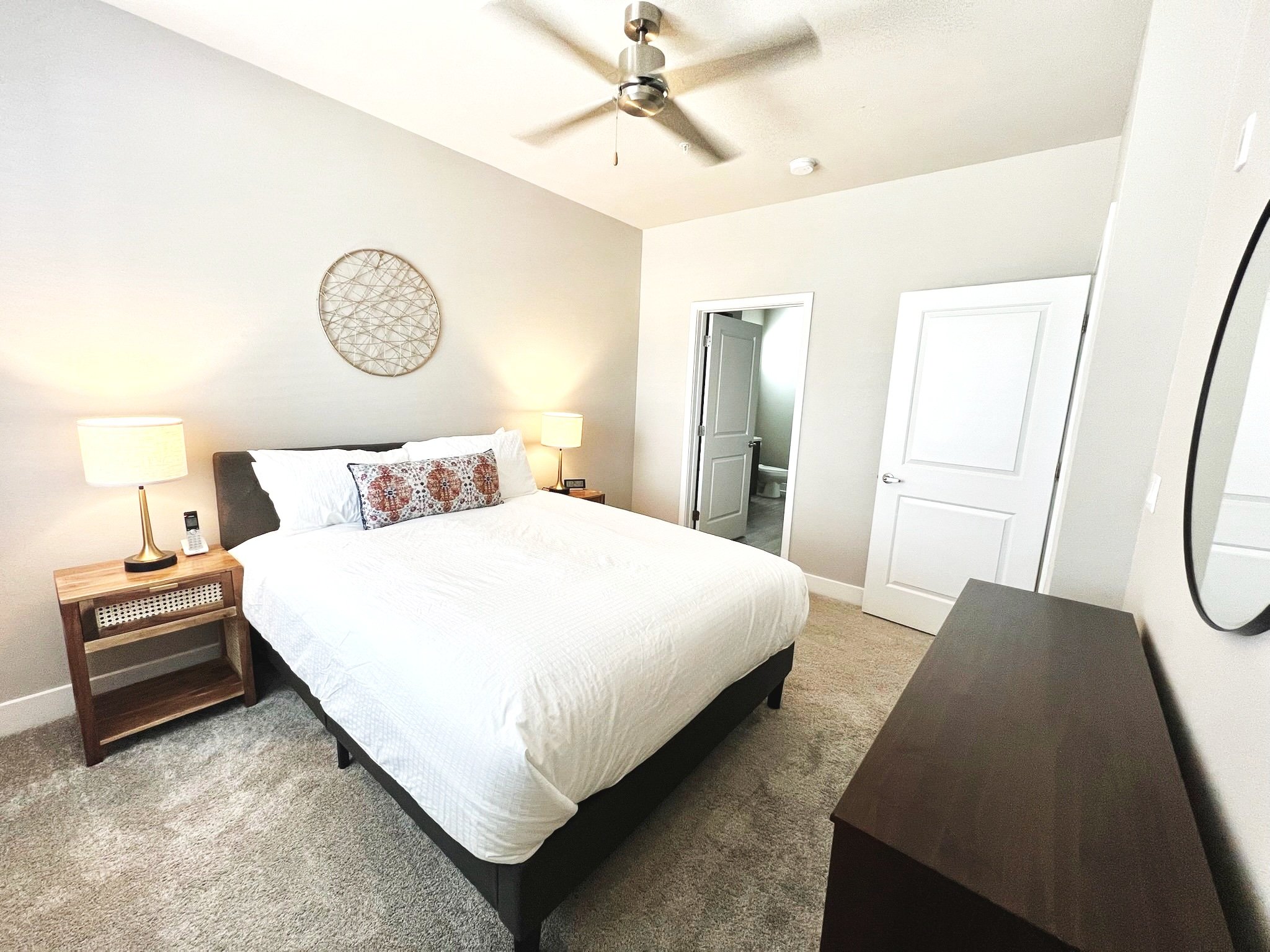 A bedroom with a white bed, two nightstands with lamps, a ceiling fan, and a decorative wall hanging, with a door leading to a bathroom.
