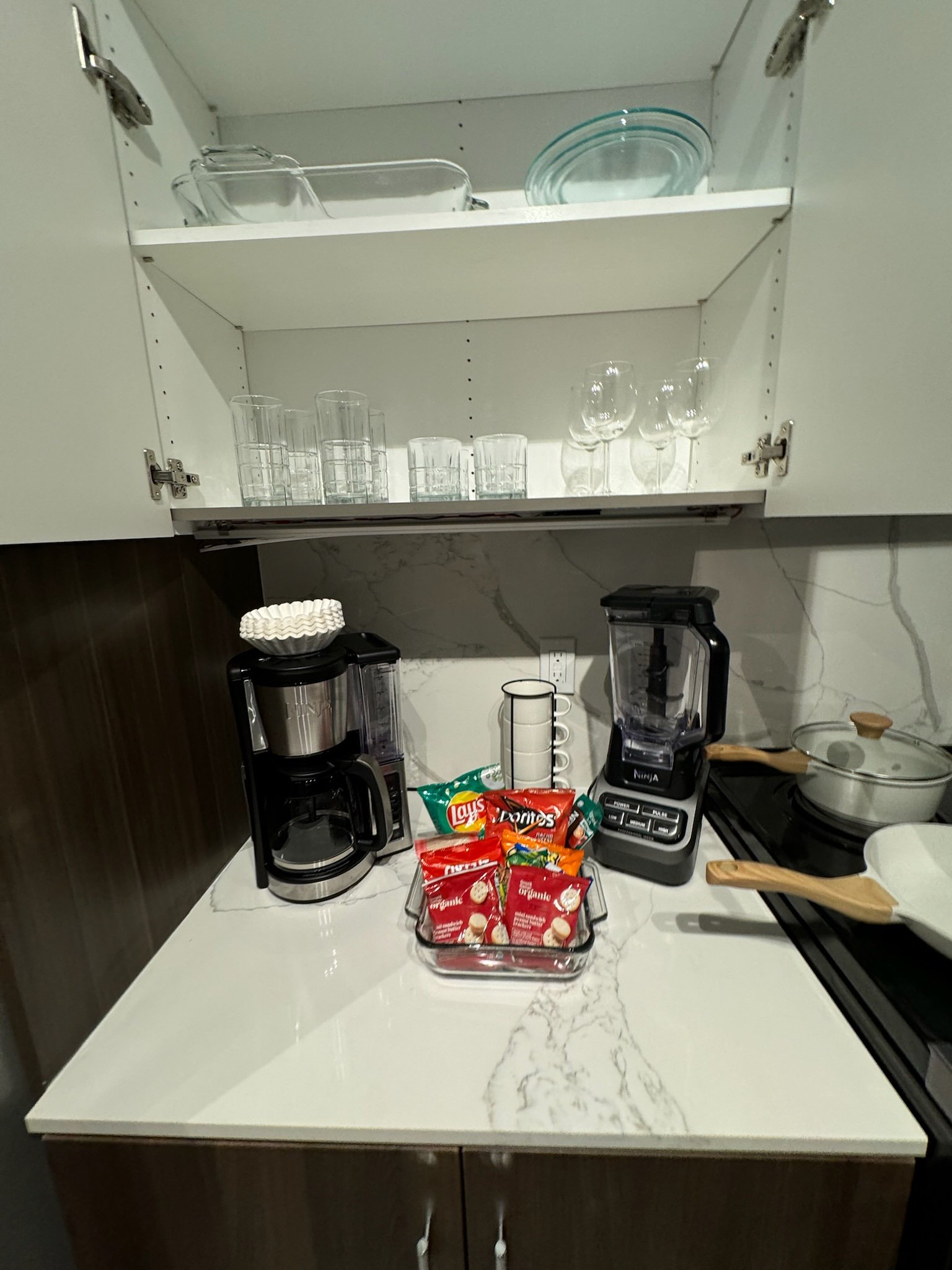 Kitchen countertop with coffee makers, a tray of red and yellow snack bags, a stack of white coffee cups, and a blender, with an open cabinet above holding glassware.