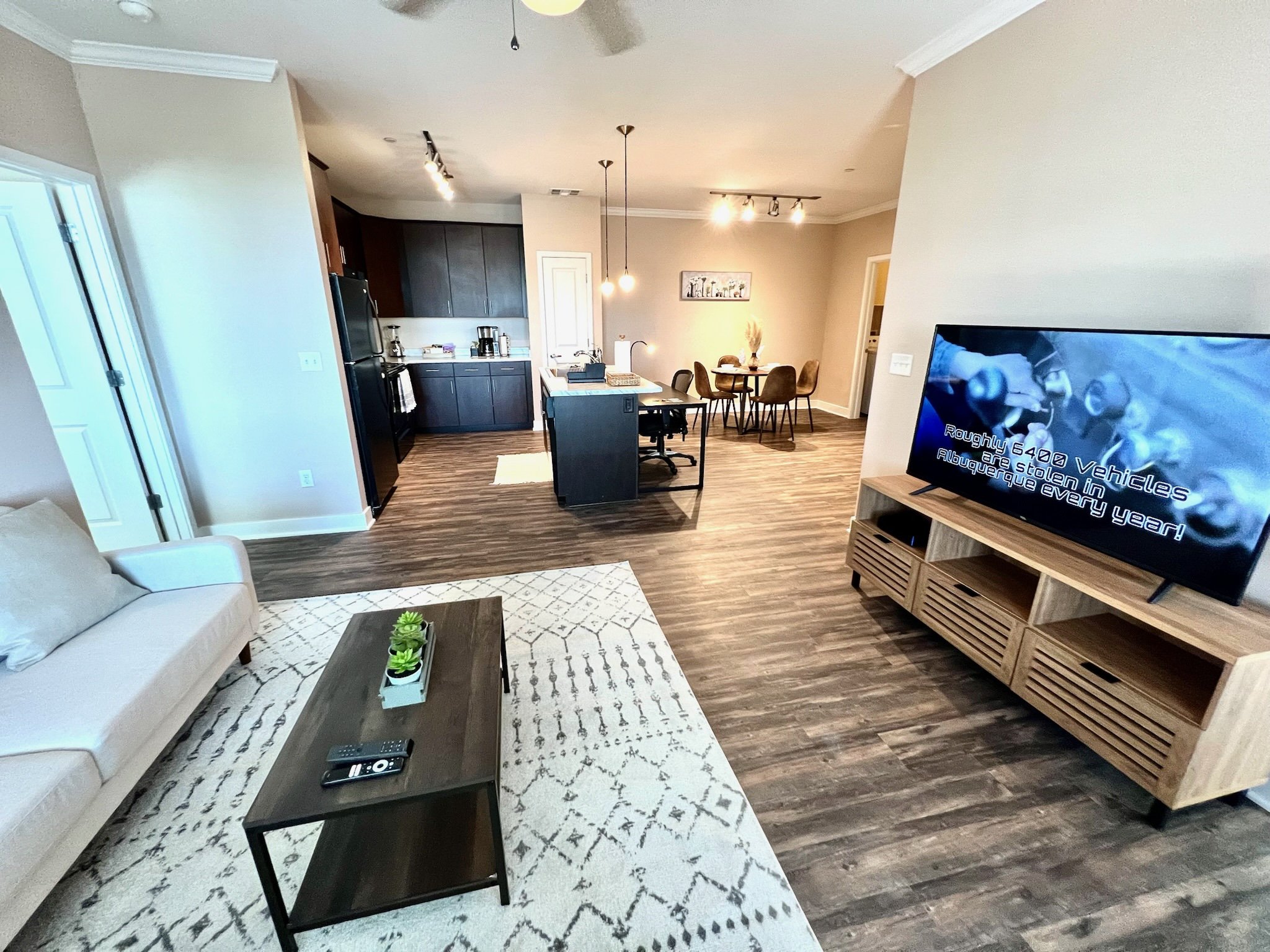 Open-concept living room and kitchen with beige walls, dark wood flooring, a white sofa, black coffee table with remote, a TV on a light wood stand, and a dining area in the background