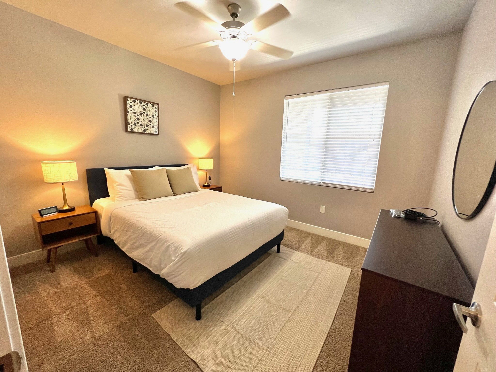 A bedroom with a bed, two nightstands with lamps, a window with blinds, a dresser with a cable and a mirror, and a ceiling fan.