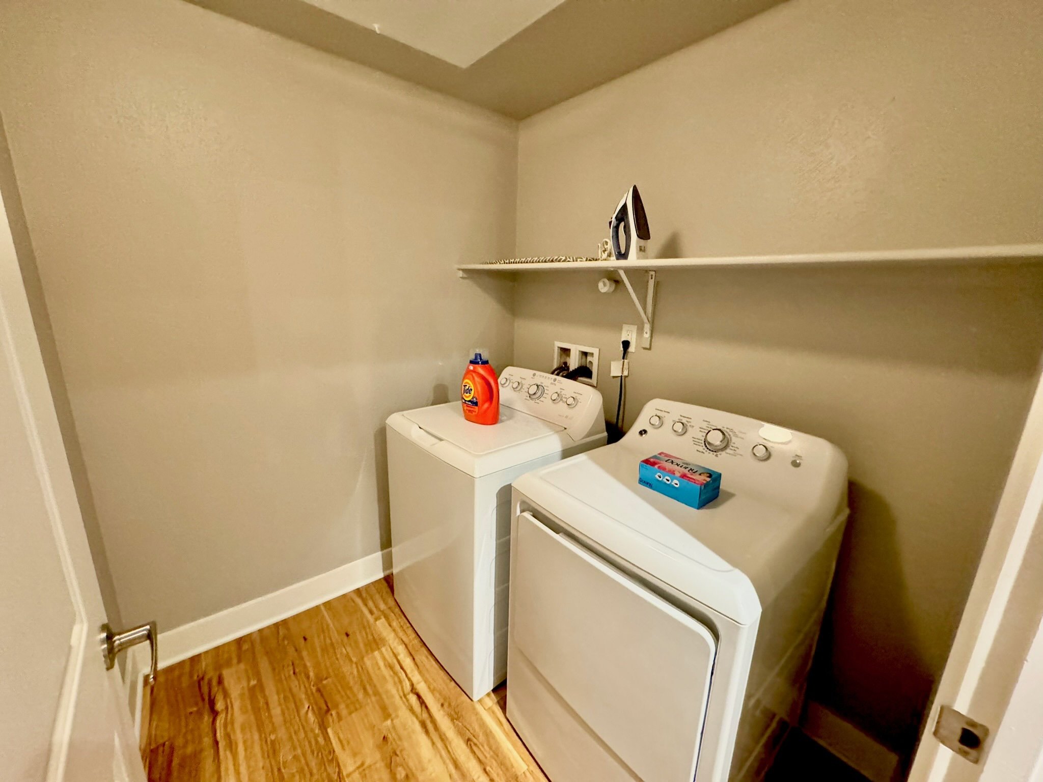 A laundry room with a washing machine and dryer, an iron on a wall shelf, and a box of dryer sheets on the dryer.
