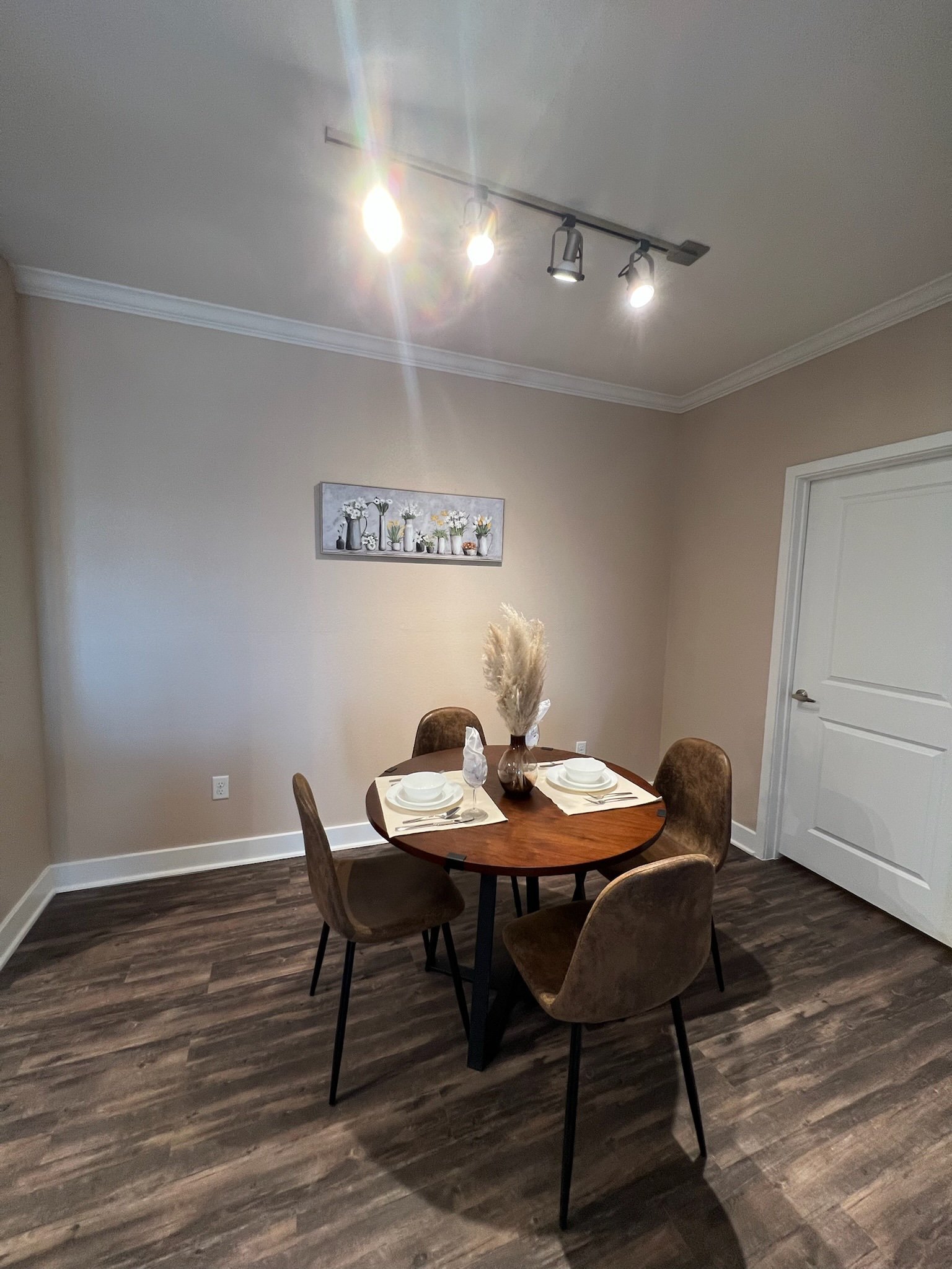 A small dining area with a round wooden table set for two, four brown upholstered chairs, a decorative vase with pampas grass, a framed flower painting, and overhead lighting in a room with beige walls and wood flooring.
