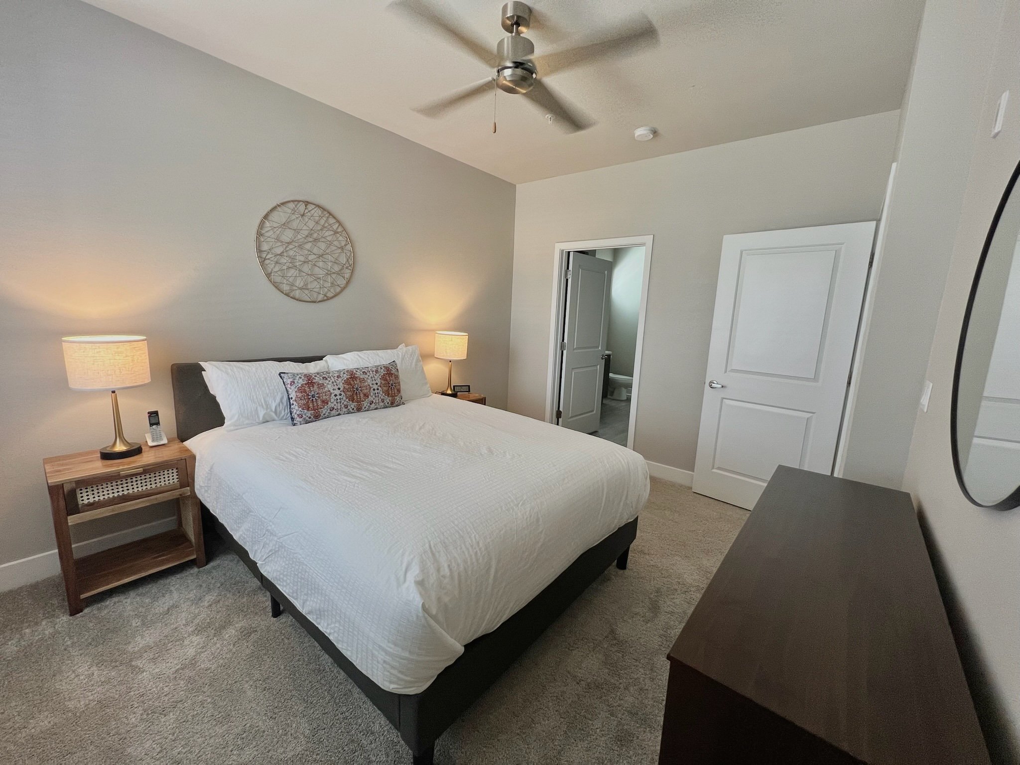 A bedroom with a bed, two nightstands with lamps, and a dresser. There is a ceiling fan and an open door leading to a bathroom.