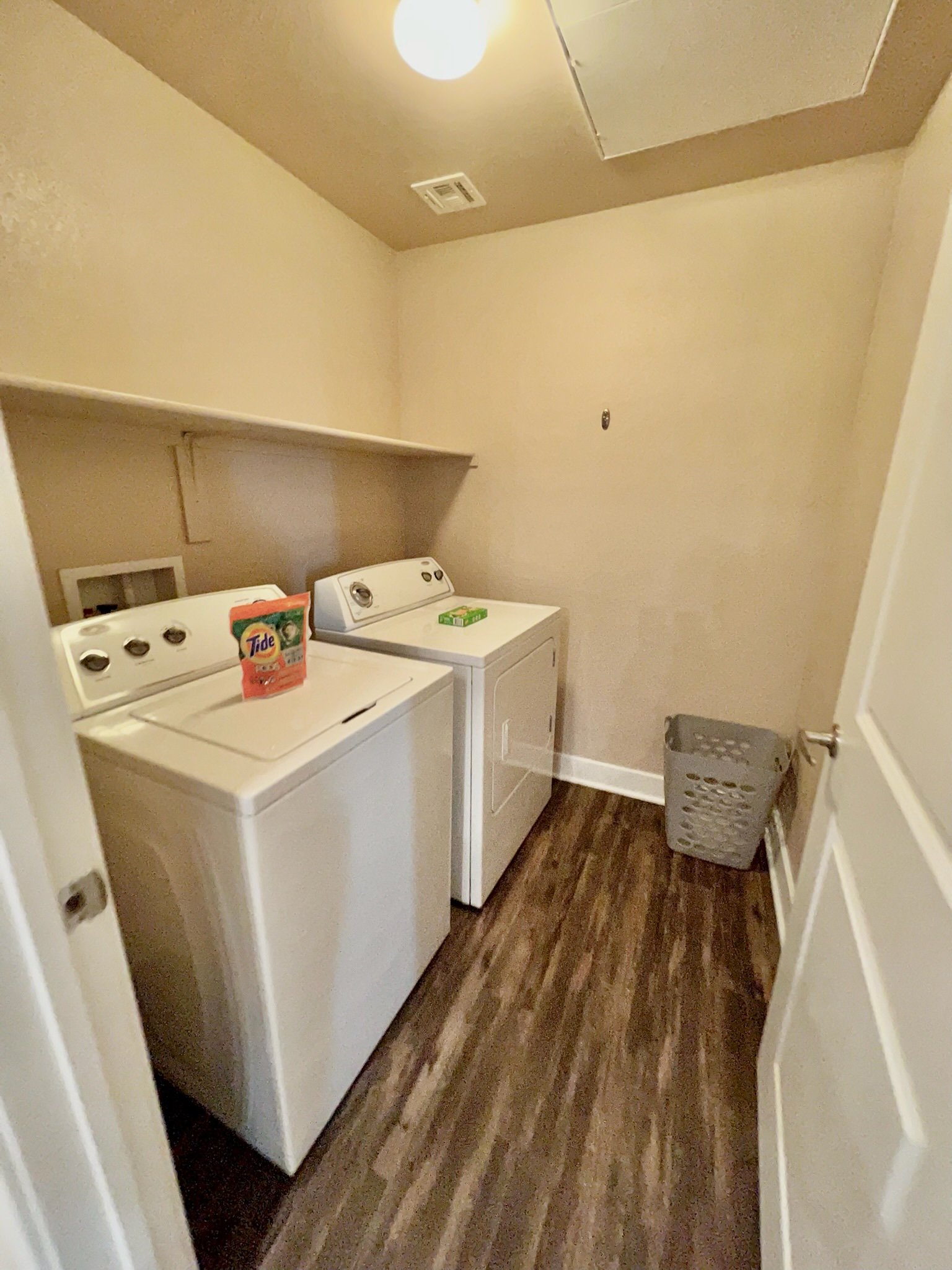 A small laundry room with a white top-loading washing machine and a white dryer next to each other. There is a shelf above the appliances and a gray laundry basket in the corner. The room has beige walls and wood-look flooring.
