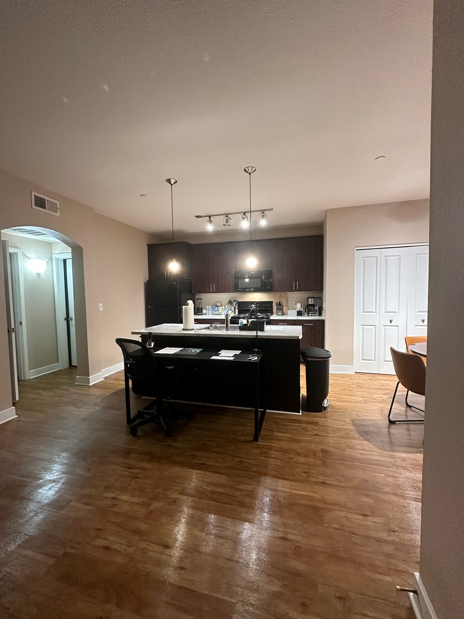 Modern kitchen with dark wood cabinets, white marble island, black appliances, and a dining area with chairs on right. Hardwood flooring and pendant lights hanging over the island.