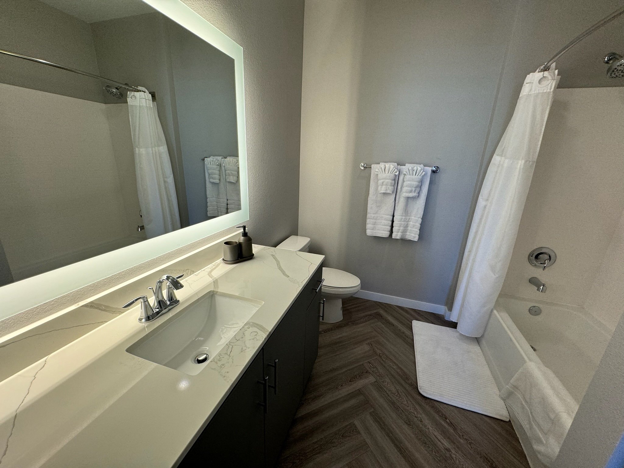 Modern bathroom with a white marble vanity, large mirror, towel rack with two white towels, toilet, bathtub with a shower curtain, and a white bath mat on wood-look flooring.