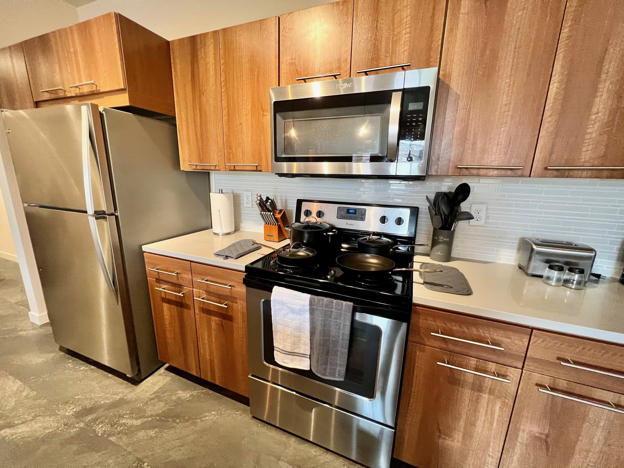 Kitchen with wood cabinets, stainless steel refrigerator, microwave, and stove with cooking utensils and towels.