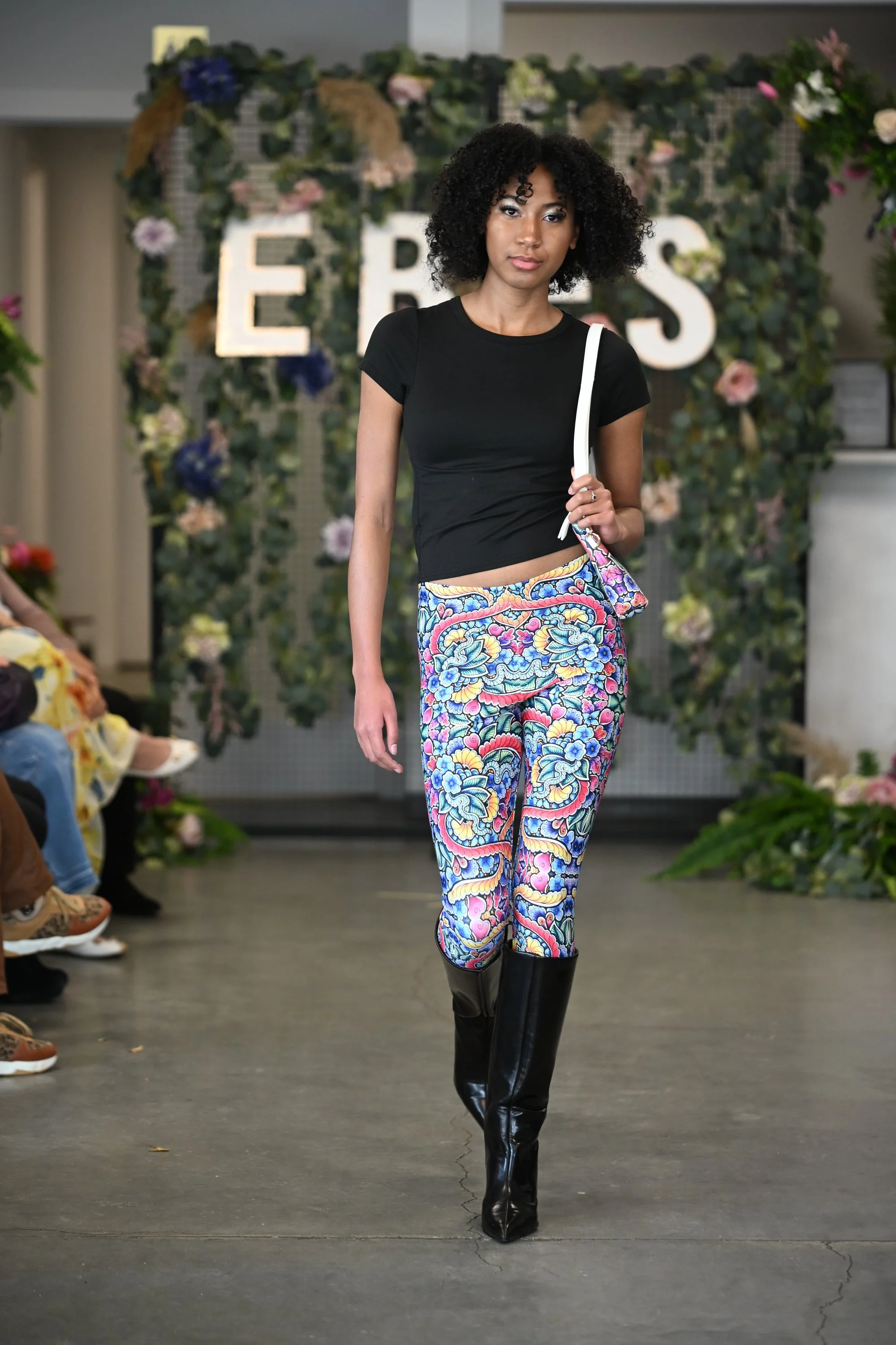 Model walking on runway wearing patterned pants based on a DoodlyDoo doodle, black top, and tall black boots with an audience watching on either side.