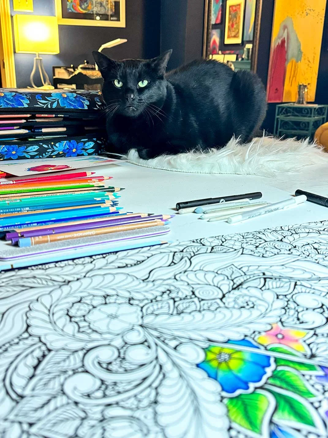 A black cat with green eyes lying on a white fur rug next to a partially colored intricate doodle on a table. The table also has colored pencils, markers, and coloring supplies.