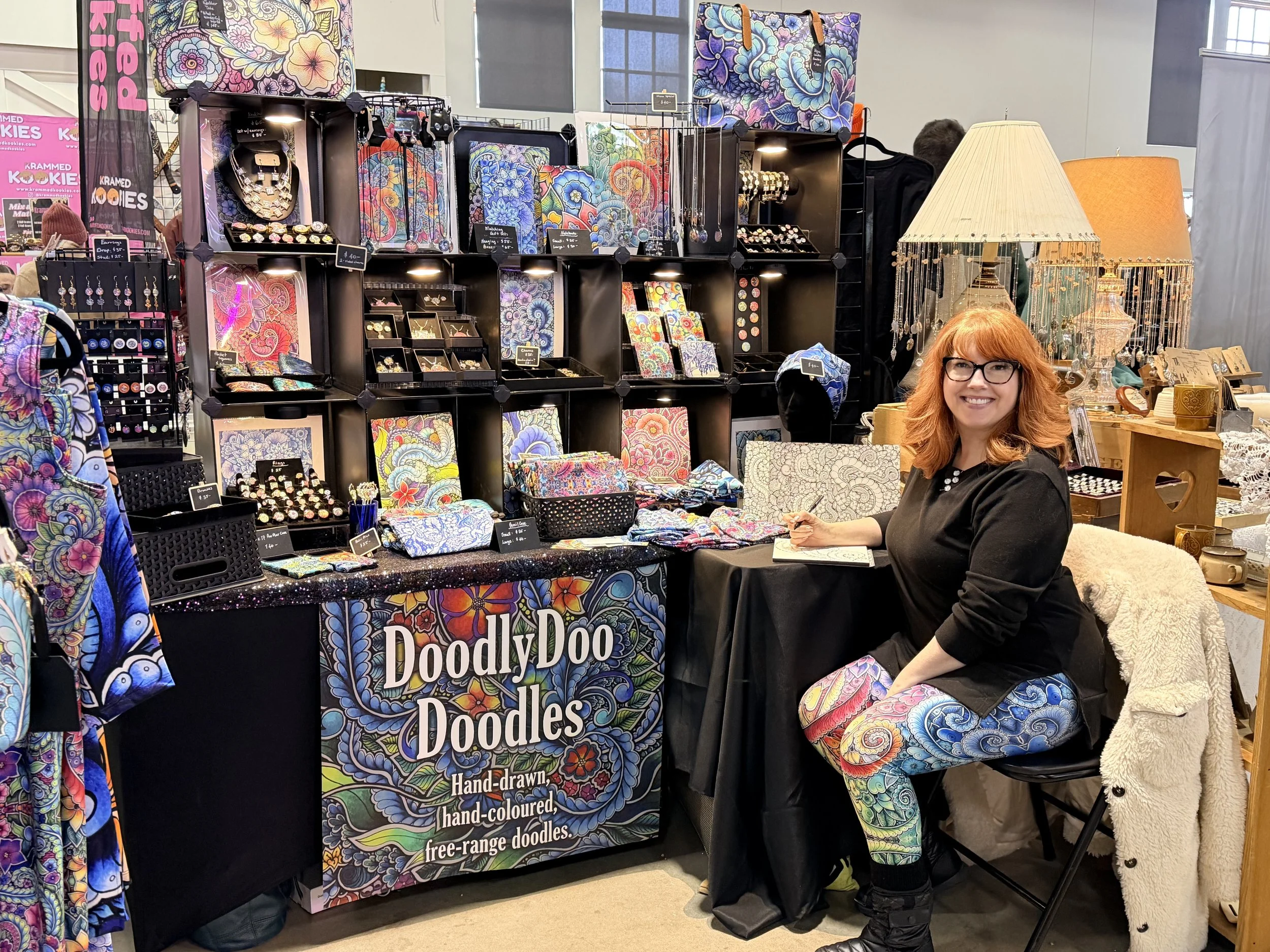 A woman with red hair and glasses sitting at a booth selling colorful, hand-drawn, and hand-colored doodle art and jewelry at a craft fair. The booth displays various art pieces, jewelry, and lamps.