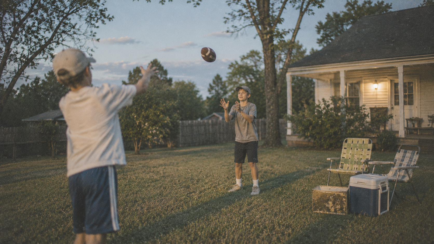 Two boys tossing a football in a backyard at dusk near a front porch, lawn chairs and a cooler set up nearby