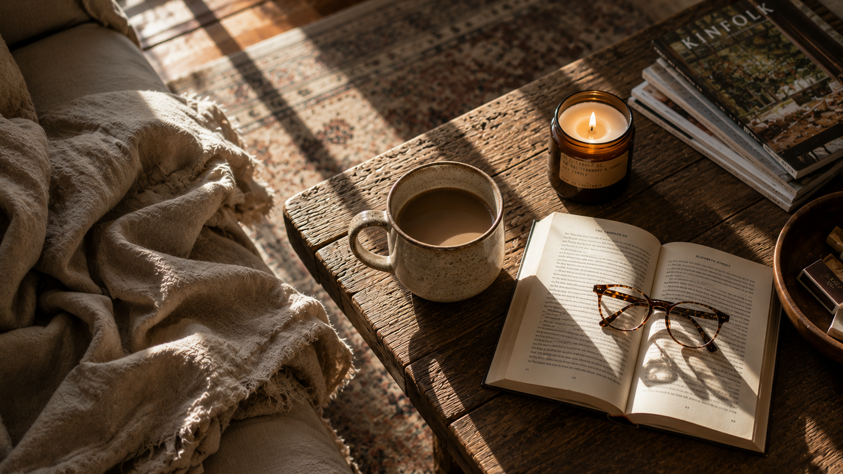 Steaming mug of coffee, lit candle, and open book with reading glasses on a rustic wooden coffee table in warm afternoon light
