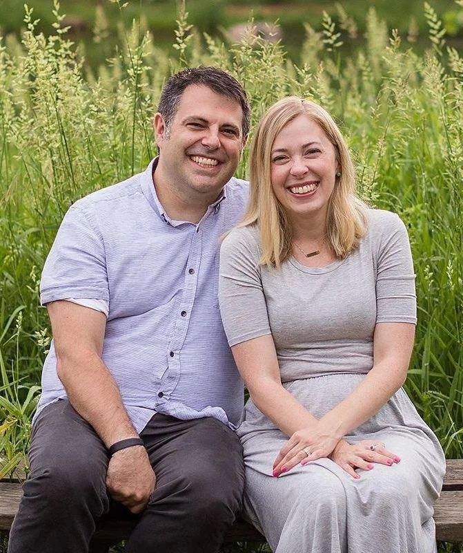 A smiling man and woman sitting on a wooden bench outdoors surrounded by tall green grass and wildflowers.