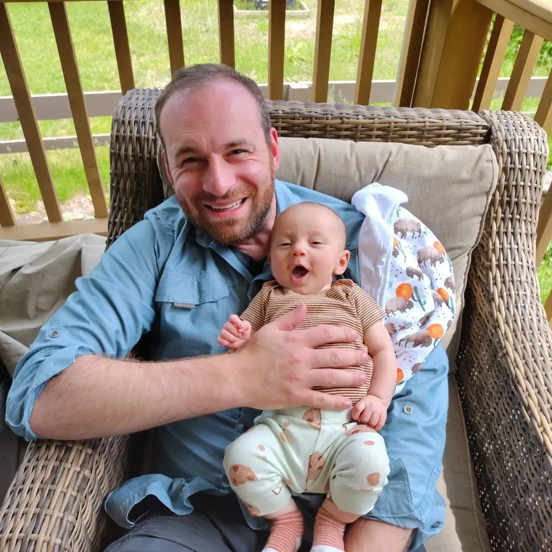 A man and a baby sit on a wicker outdoor couch on a porch with wooden railings, with greenery in the background. The man is smiling and holding the baby, who appears to be yawning or making a face.