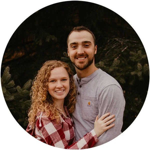 A smiling couple outdoors in front of evergreen trees, the woman has curly red hair and wears a red plaid shirt, while the man has short dark hair and wears a light gray shirt.