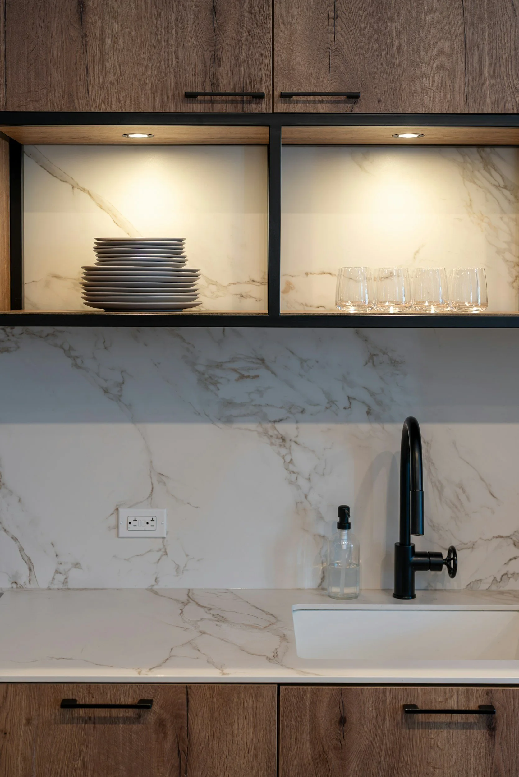 Modern kitchen with wooden cabinets, marble backsplash, black faucet, stacked white plates, clear glasses, and a soap dispenser. Rhinebeck home management property care.