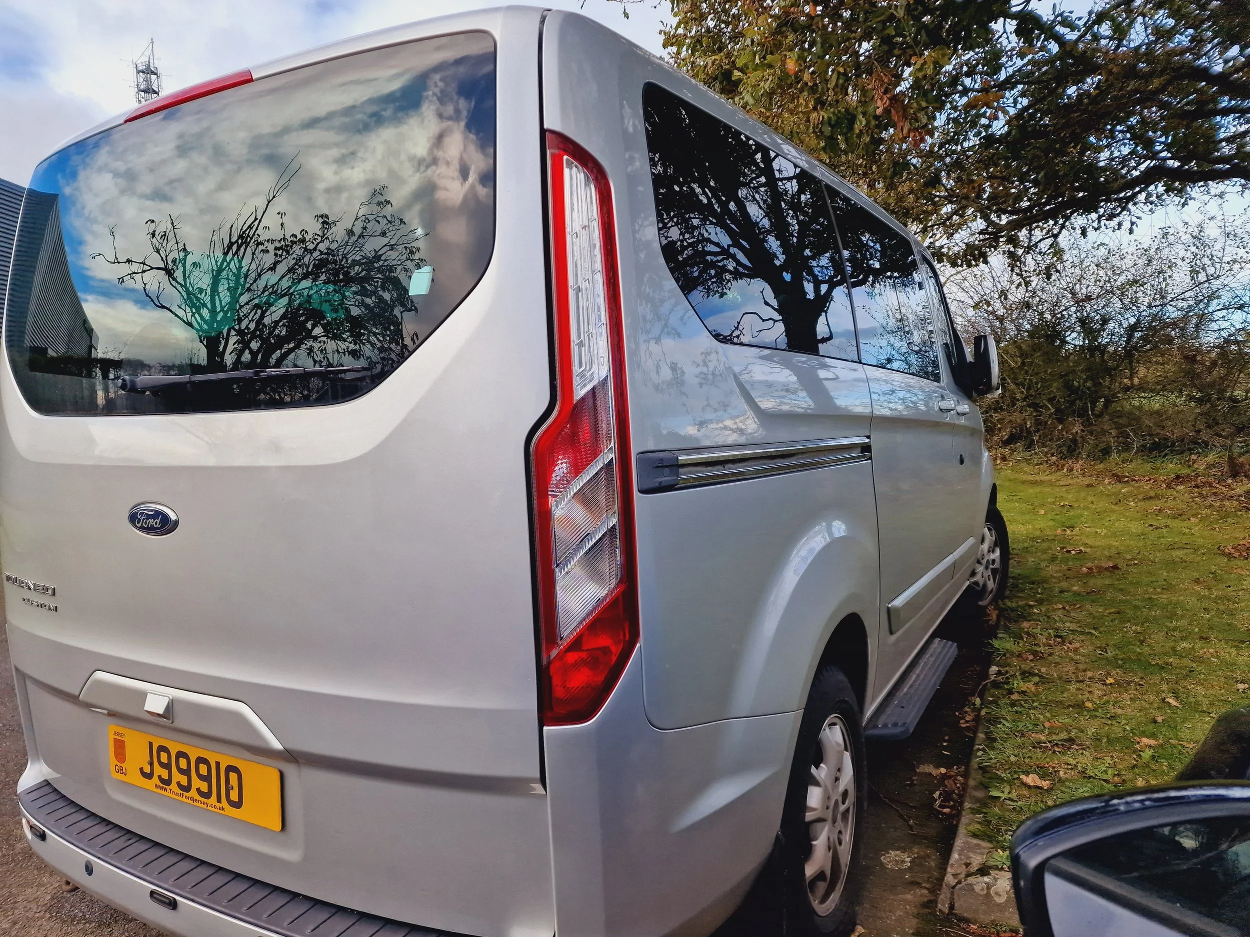 A silver Ford Transit van parked on a grassy area next to a sidewalk with trees, reflecting clouds and sky in the rear window.