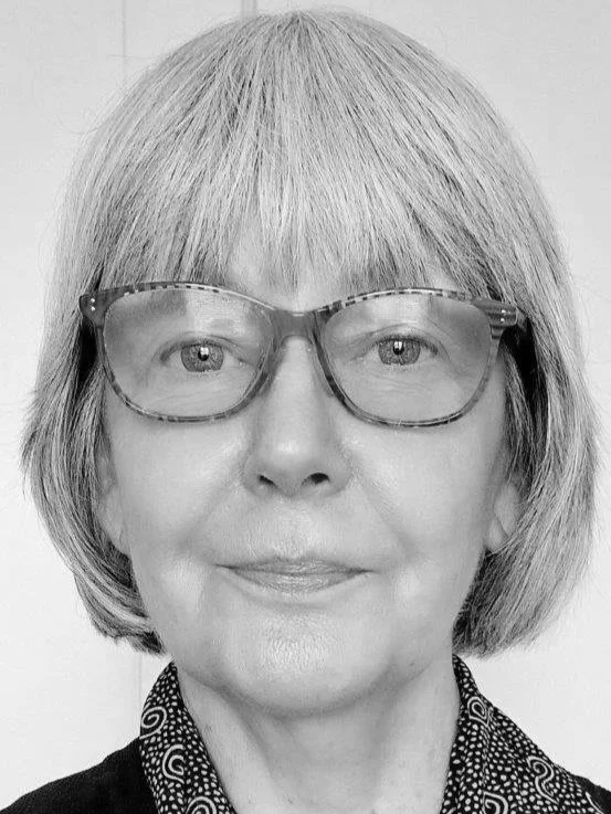 Close-up of an older woman with gray hair and colorful glasses, smiling lightly, wearing a black shirt with a patterned collar, standing against a light-colored background.