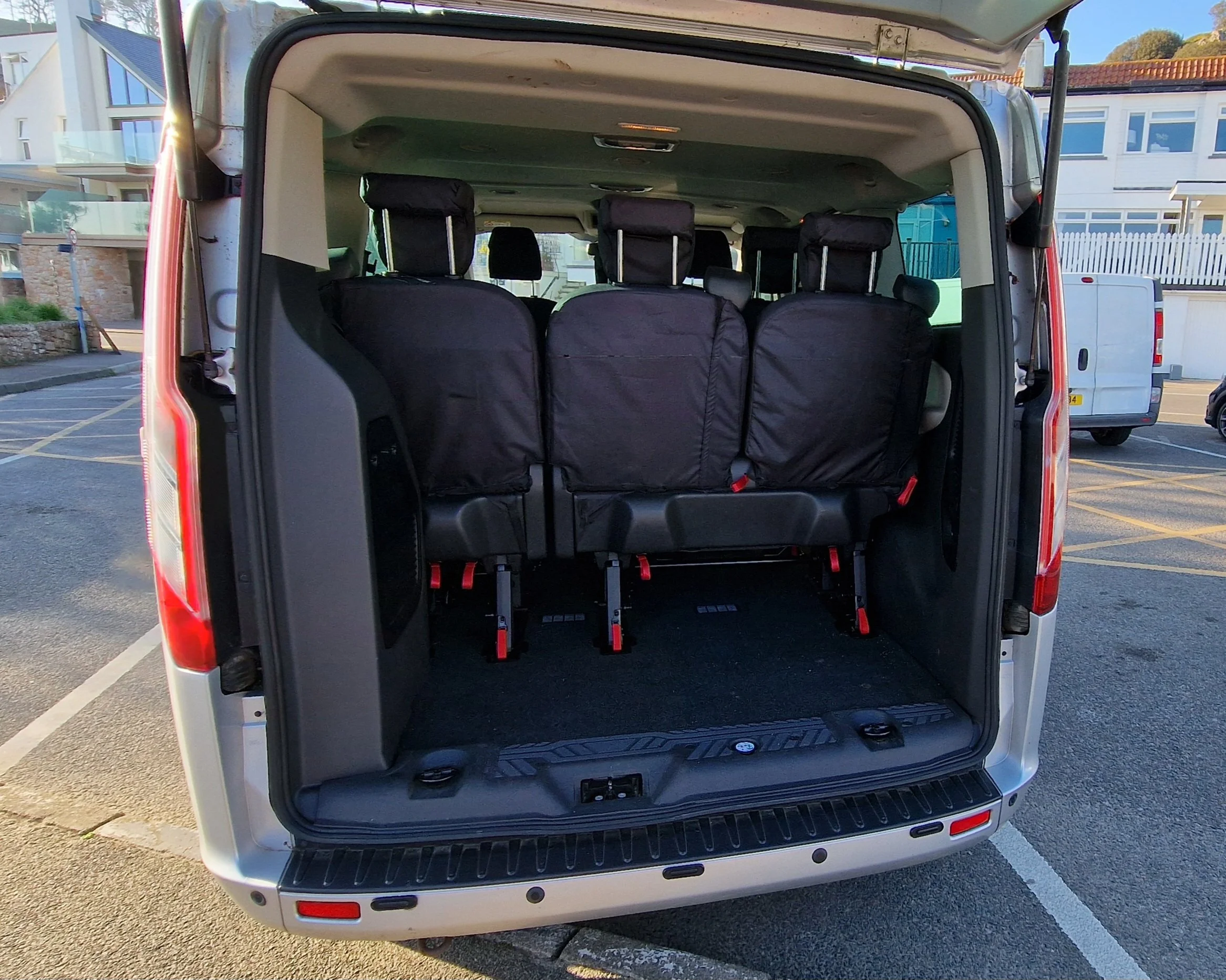 Rear view of a van with the back doors open, showing three rows of black seats, with some seats covered in black protective covers, parked in a parking lot in front of modern buildings.