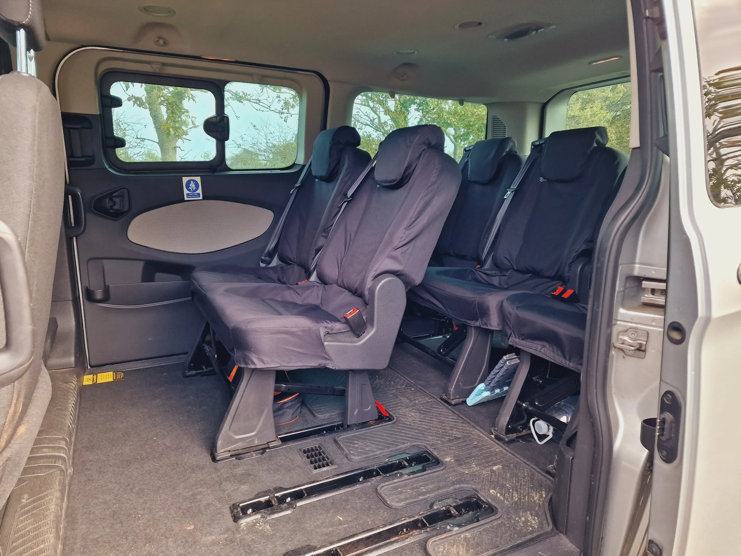 Interior of a passenger van showing multiple black fabric seats with headrests, windows with greenery outside, and a sliding door on the right side.