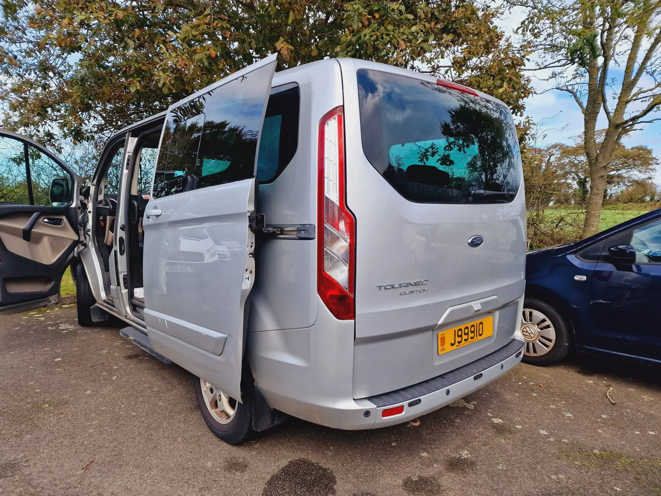 Silver Ford Tourneo Custom shuttle van with an open front door, parked on an outdoor lot with trees in the background.