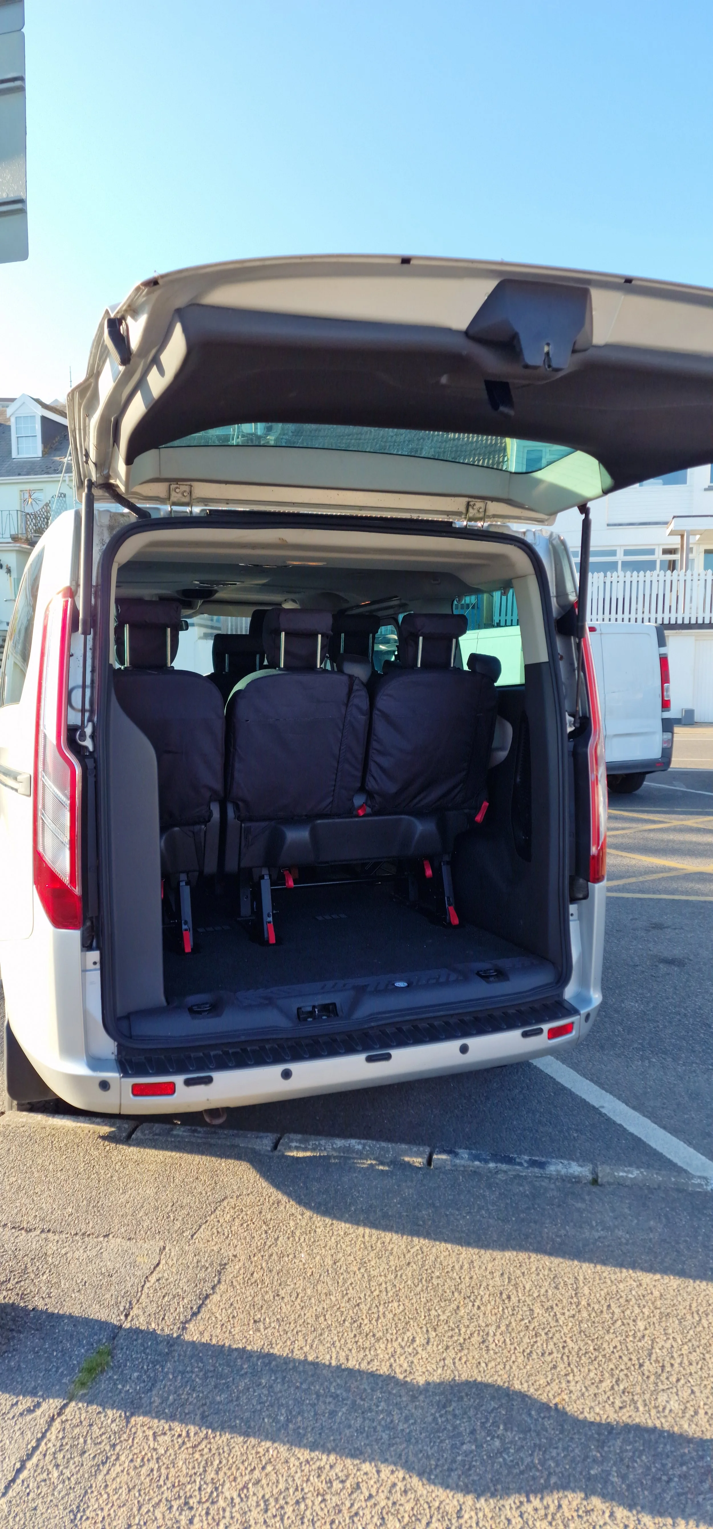 Open rear view of a silver minivan with three black-folded seats inside, parked in a parking lot with other vehicles and buildings in the background.