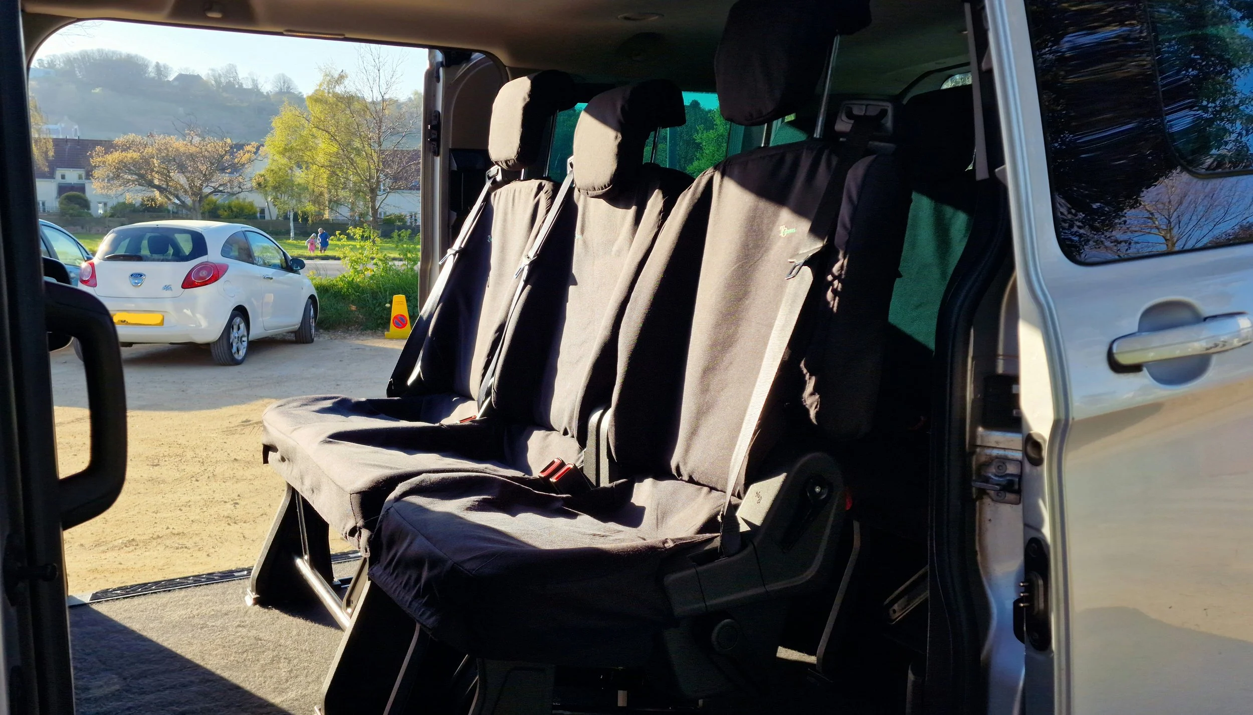 Inside a vehicle showing three black fabric seats with headrests, with an open door revealing the exterior parking lot.