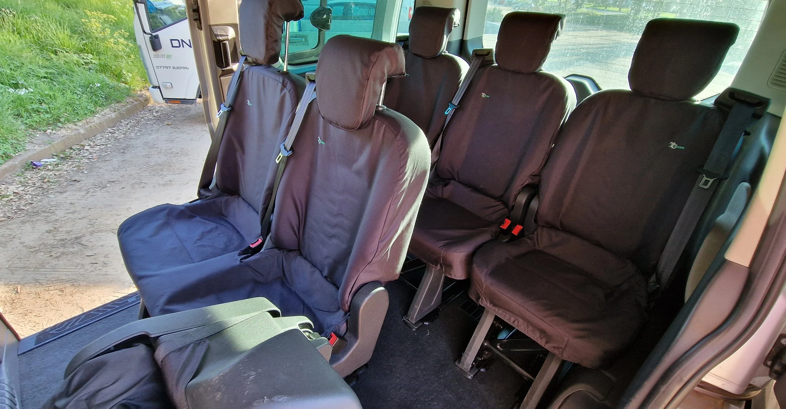 The interior of a passenger bus showing two rows of black fabric seats with headrests, some with seatbelt straps, and the bus door open revealing an outdoor scene with grassy area and dirt ground.