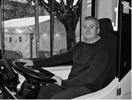 A man sitting in the driver's seat of a bus or large vehicle, smiling at the camera.