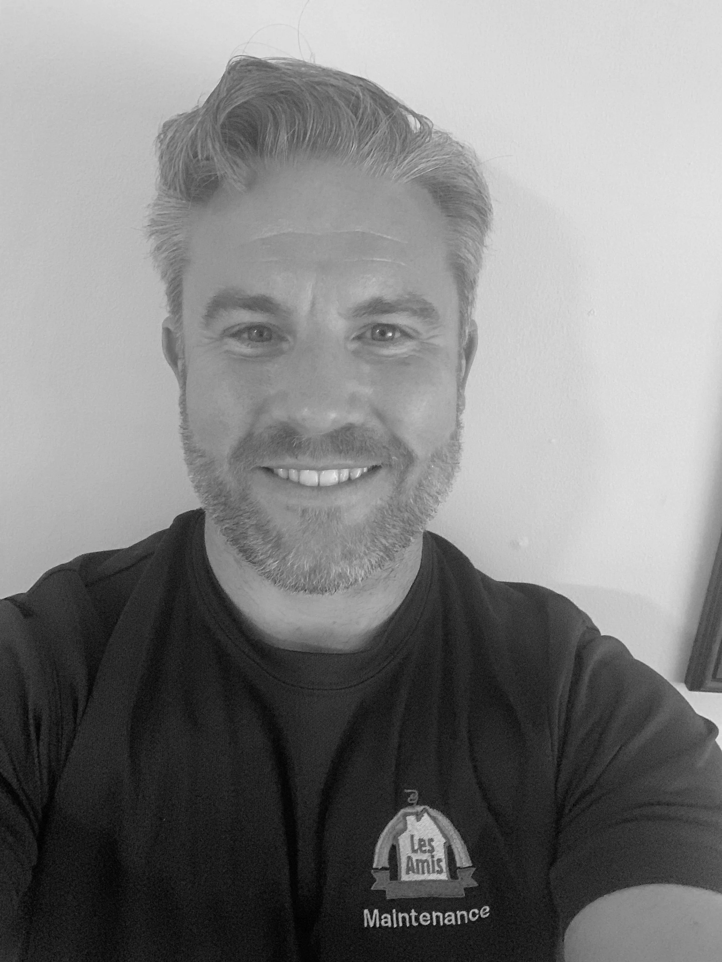 A smiling man with salt and pepper hair and beard, wearing a black shirt with a logo that reads 'Les Amis Maintenance,' taking a selfie in front of a plain beige wall.