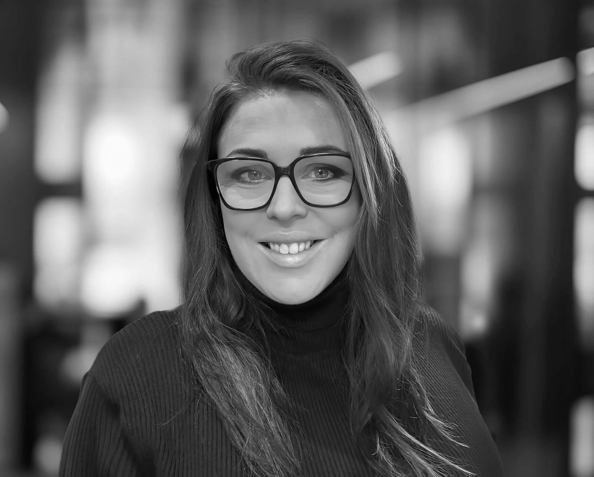A woman with long brown hair, wearing black glasses and a black turtleneck sweater, smiling in an indoor setting with blurred background.