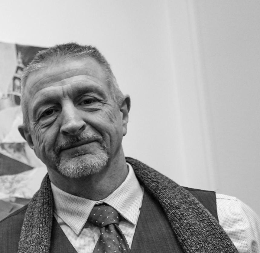A middle-aged man with short gray hair and a gray beard, wearing a suit, purple tie, white shirt, and gray scarf, smiling at the camera.