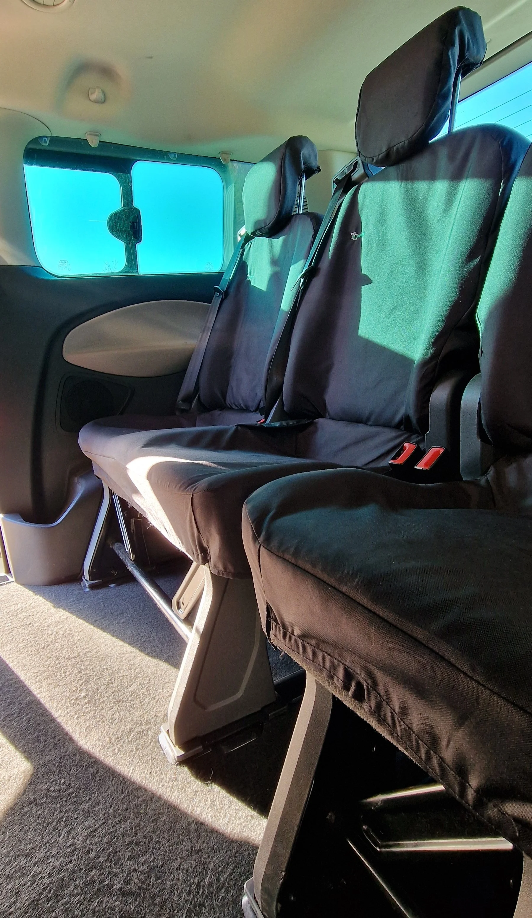 Inside a vehicle showing three black seats with headrests, armrests, and seat belts. The window reveals a clear blue sky outside.