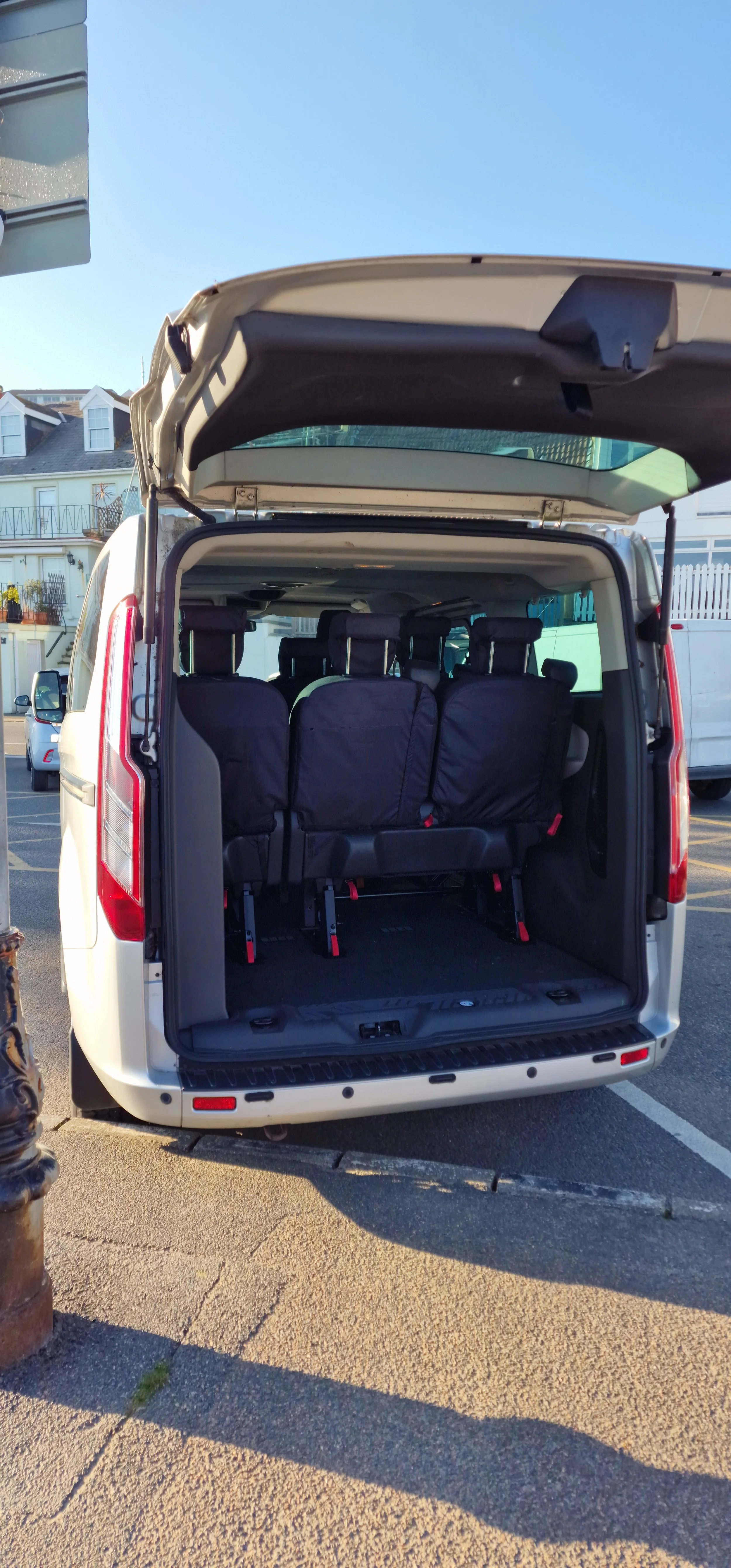 Back of a white van with open rear door, showing three folded black seats inside, parked in a lot with residential buildings in the background.