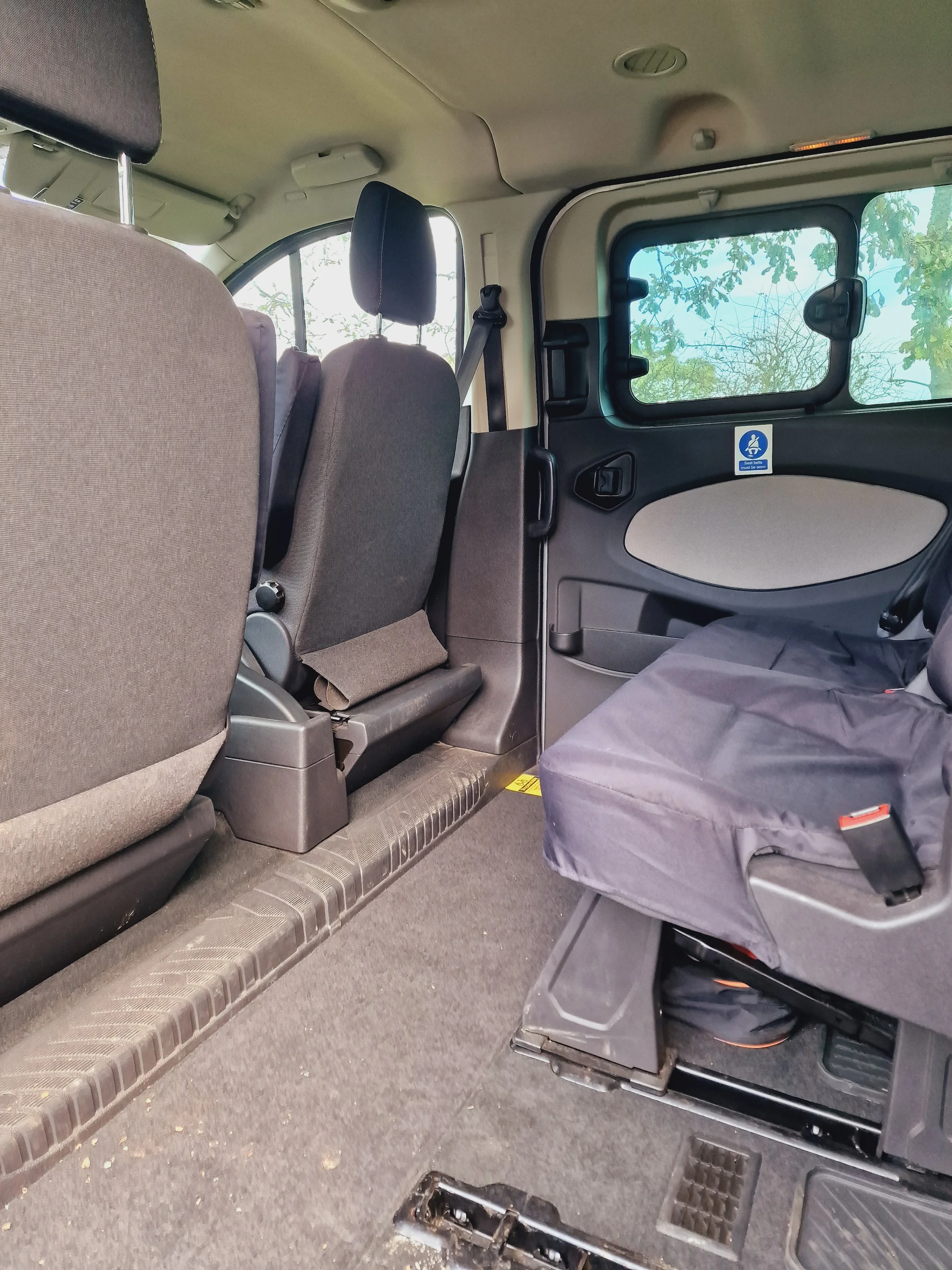 Inside the rear section of a vehicle with two row seats, a child's car seat, and a window showing trees outside.