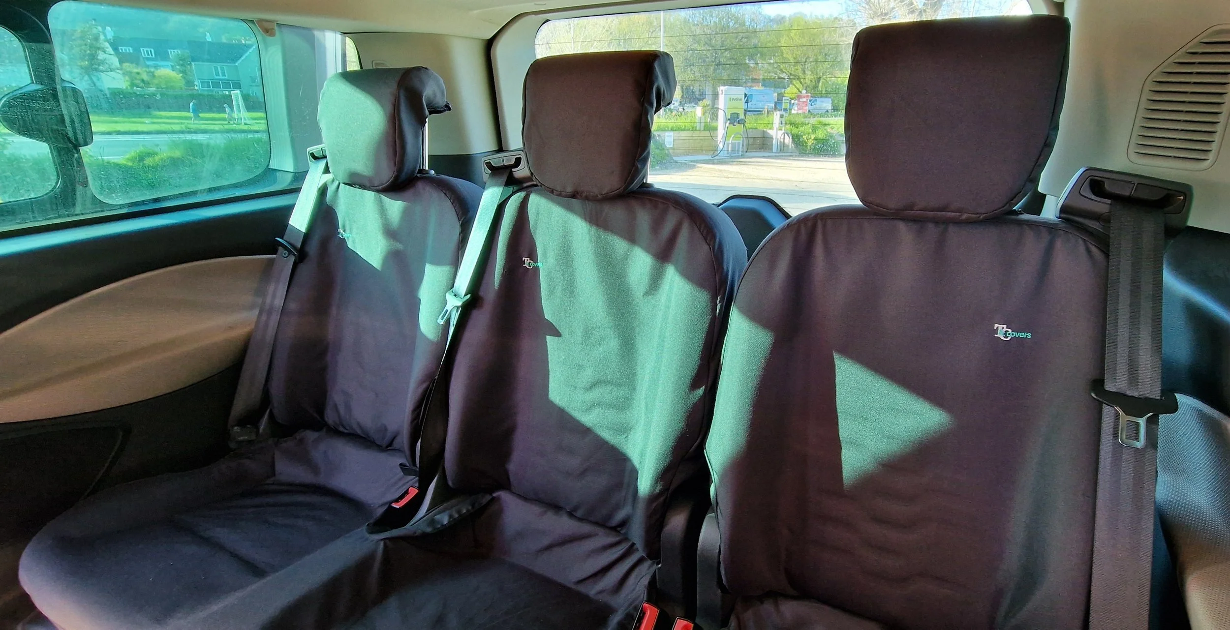 The image shows the interior of a vehicle with three black seats, all equipped with headrests and seatbelts. The seats are covered with protective seat covers. Outside the vehicle window, there is a view of a parking lot with a charging station and s