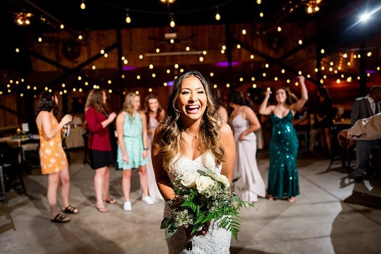 Bride holding a bouquet, smiling, surrounded by female guests at a rustic wedding reception 