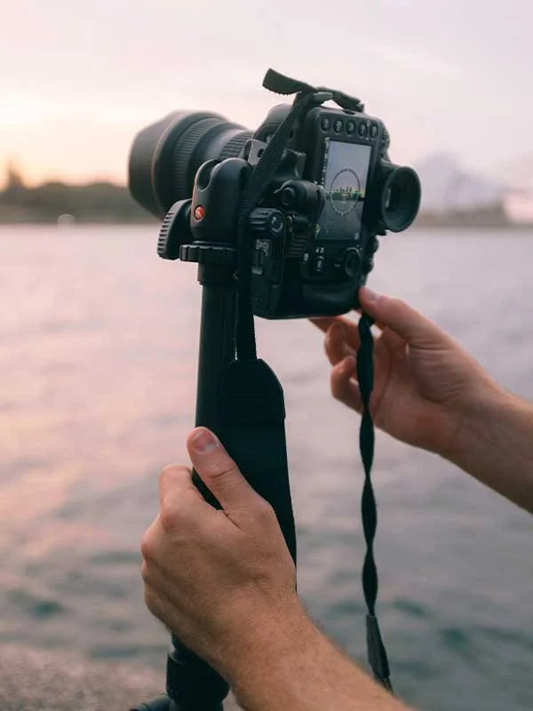 Person holding a DSLR camera on a monopod near a waterfront at sunset.