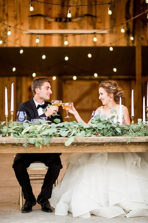 Bride and groom toasting at a rustic wedding reception table decorated with greenery and candles
