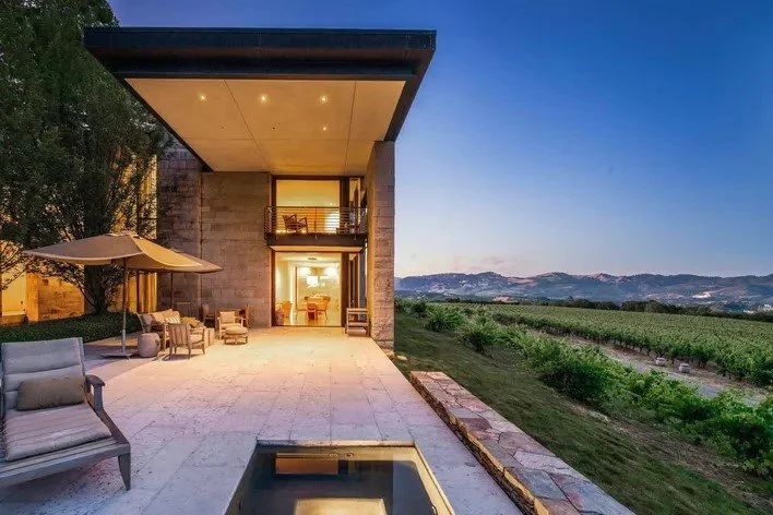 Modern two-story house with large patio, outdoor seating, and a small pool, overlooking a vineyard and distant mountains at sunset.