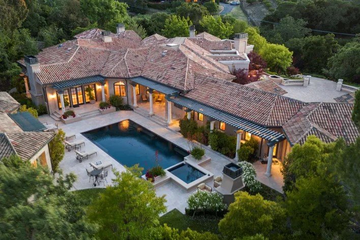 A spacious backyard with a swimming pool, hot tub, and patio area surrounded by a large, luxury house with multiple roofs and outdoor lighting, set amidst lush greenery.