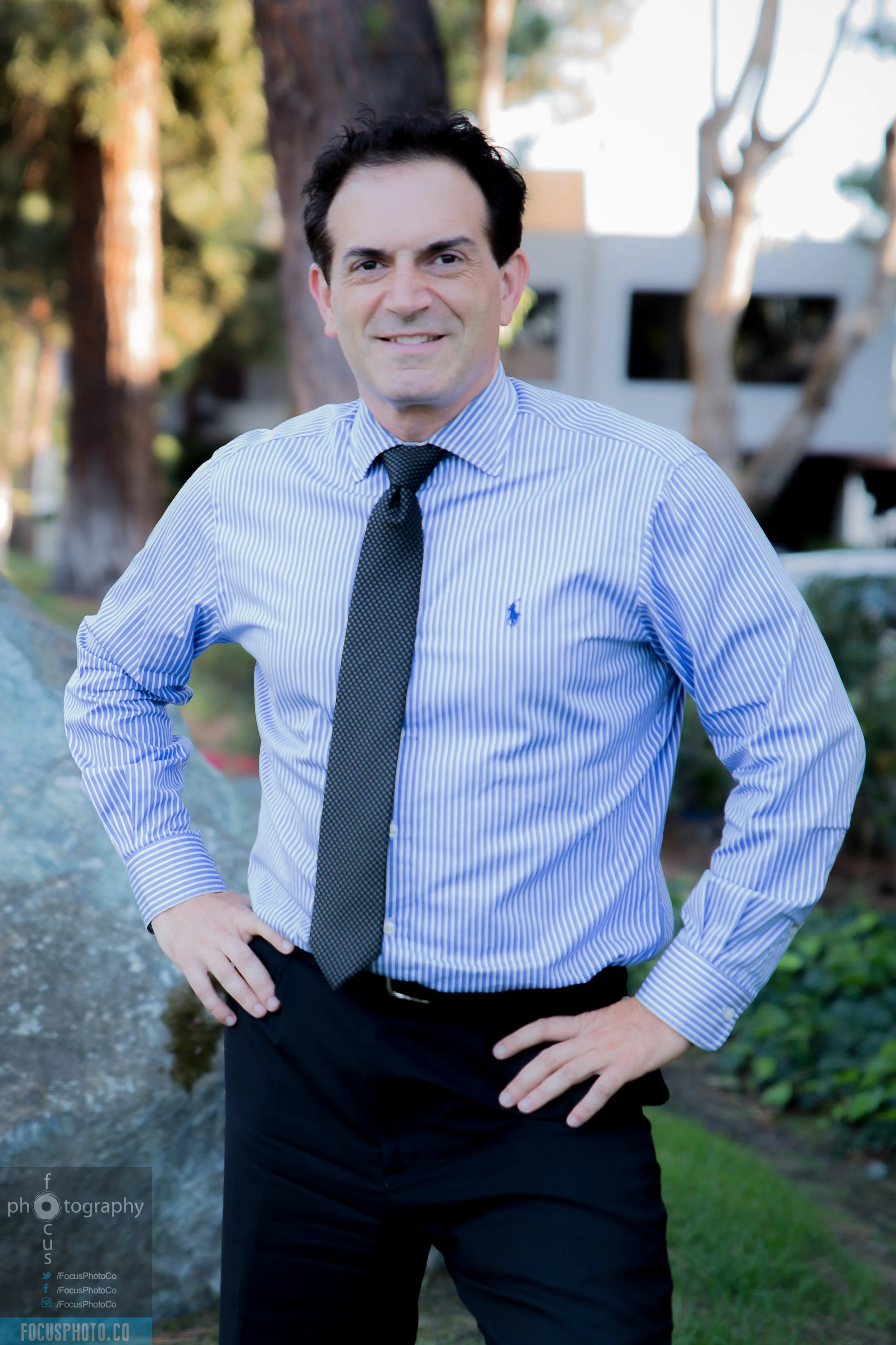 A man in a blue-striped dress shirt and black tie standing outdoors with hands on hips, smiling, with trees and a building in the background. Cyrus Ghazvini, the trusted realtor to get you your dream house.