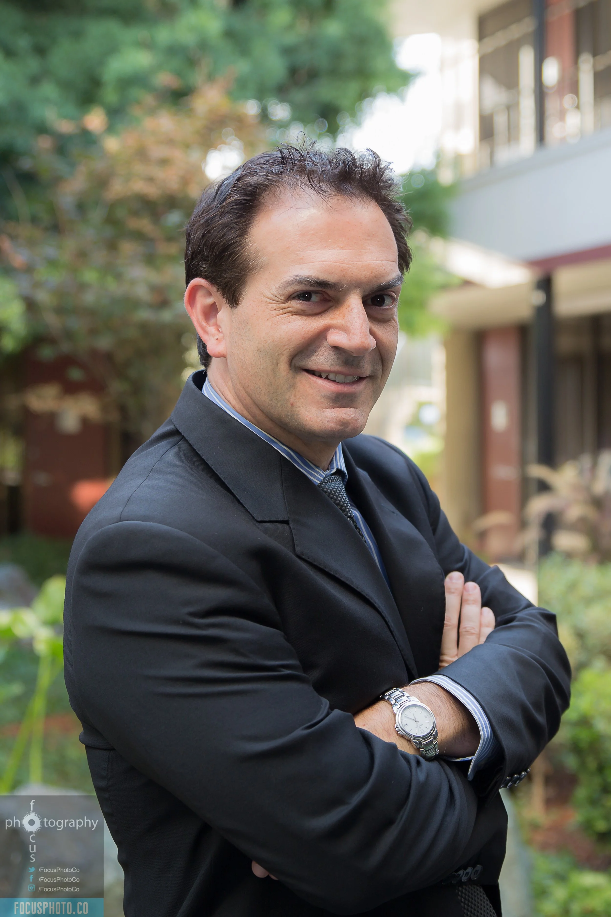 A man in a black suit with arms crossed, smiling outdoors with trees and a building in the background. Cyrus Ghazvini, the trusted realtor to get you your dream house.