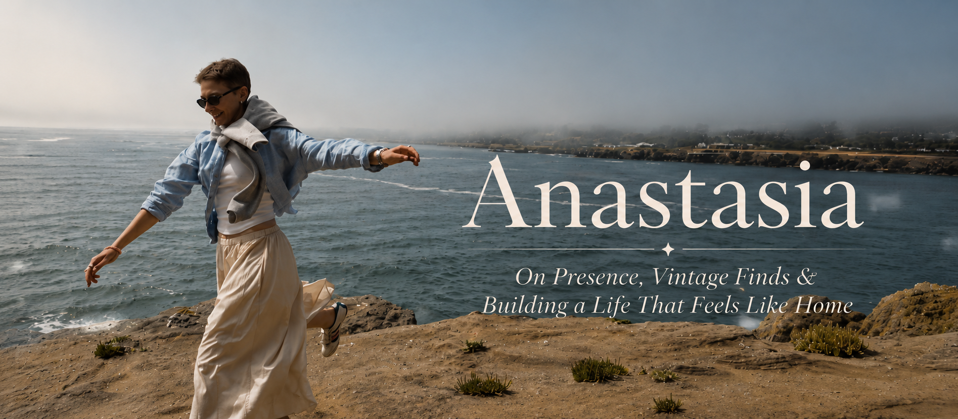 Woman in light-colored pants, white shirt, and denim jacket, smiling and jumping on rocky terrain by the ocean, with coastal landscape in the background, overlaid with text 'Anastasia' and the tagline 'On Presence, Vintage Finds & Building a Life That Feels Like Home'.