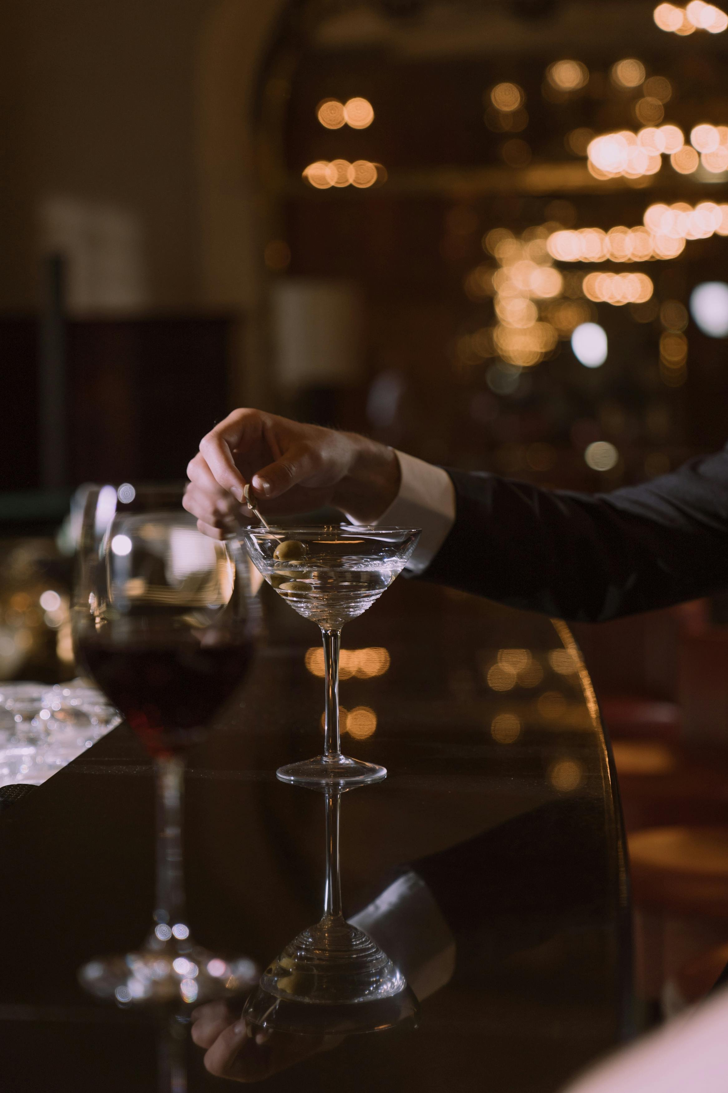 Person in suit holding a toothpick with olive over a martini glass on a dimly lit bar with reflections of lights.